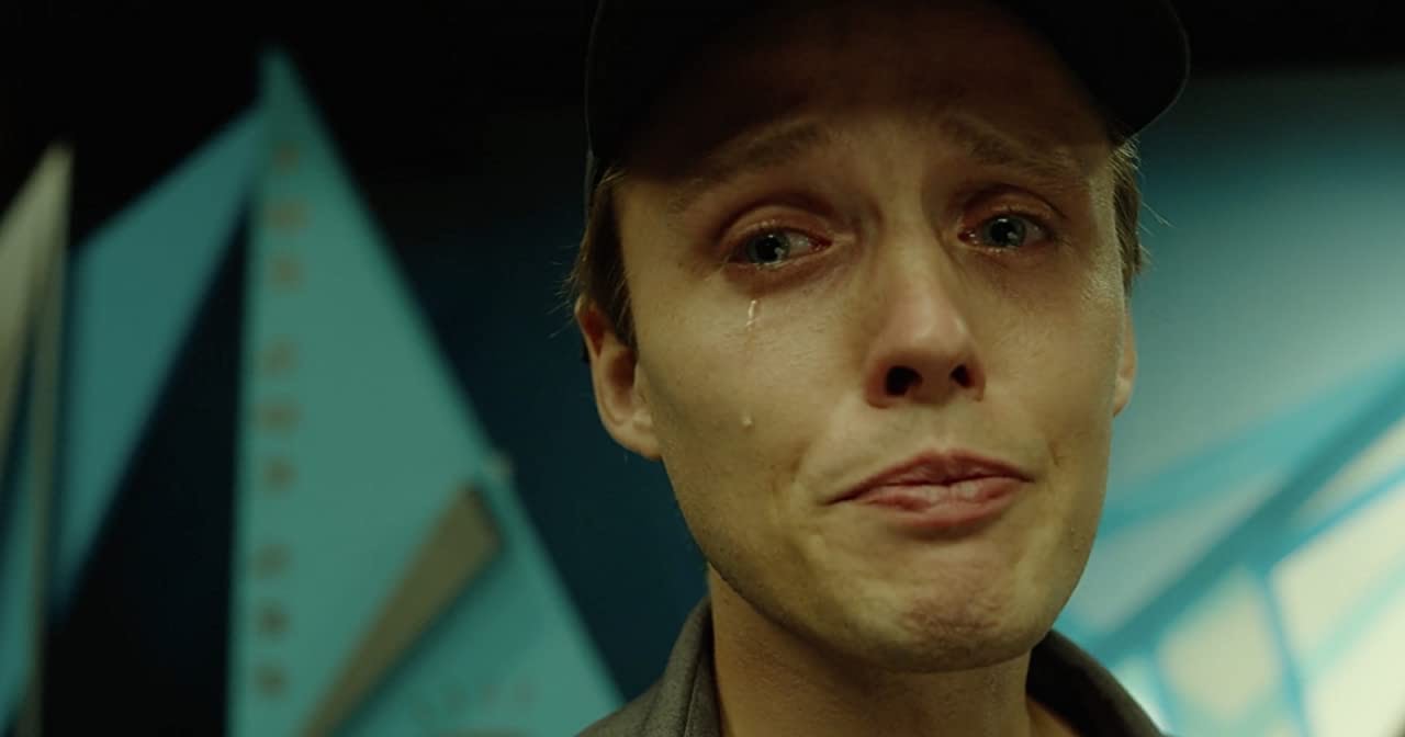 Cena do filme Interrompemos a Programação. A imagem foca o rosto do personagem Sebastian, um homem branco, loiro e olhos azuis. Na cena, o homem usa um boné preto e está chorando.