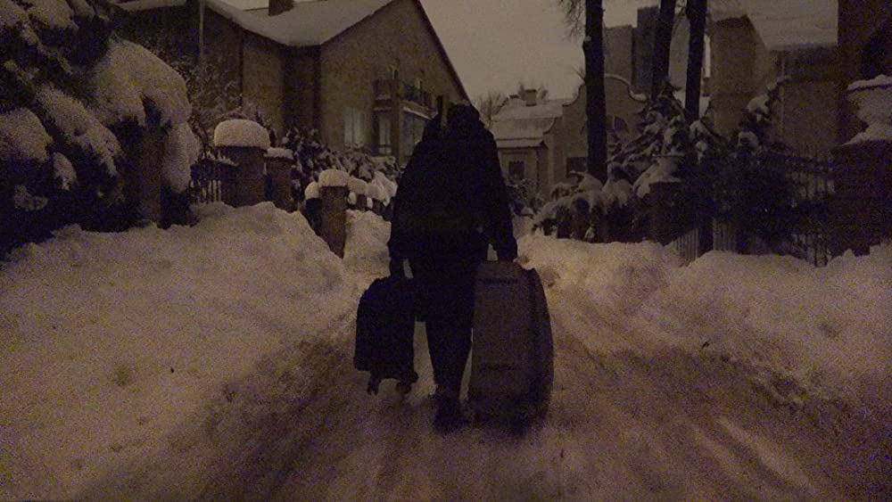 Cena do filme Bem-vindo à Chechênia exibe uma pessoa de costas, usando casaco e calça preta e arrastando duas malas. O cenário é de casas e algumas árvores cobertas de neve.
