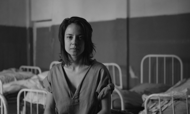 Cena da série Colônia. Na fotografia a personagem Valeska está sentada em uma das camas do hospício, a personagem veste a camisola cinza e a imagem é em preto e branco. 
