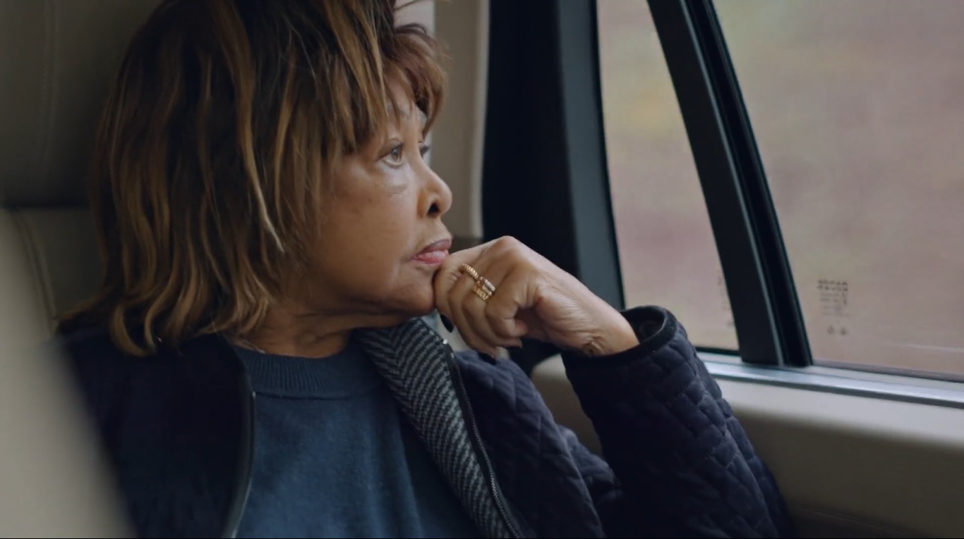 Cena do documentário Tina. Na imagem, vemos a cantora Tina Turner dentro de um carro, disposta de perfil, olhando pela janela e apoiando o rosto em sua mão fechada. Ela é uma mulher negra, de cabelos loiros, com semblante reflexivo e veste roupas de frio. 