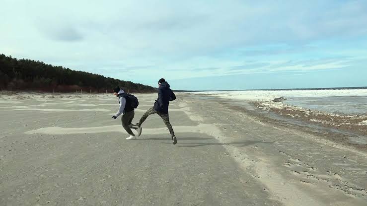 Cena do filme Bem-vindo à Chechênia em que dois homens, usando casaco e calça, se divertem em uma praia deserta. 