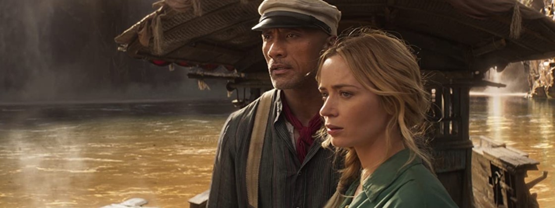 Cena do filme Jungle Cruise exibe uma mulher branca e um homem samoano em cima de um barco num rio. A mulher tem cabelo loiro longo e veste uma camisa verde. O homem usa uma boina e veste uma camisa cinza com suspensórios. 