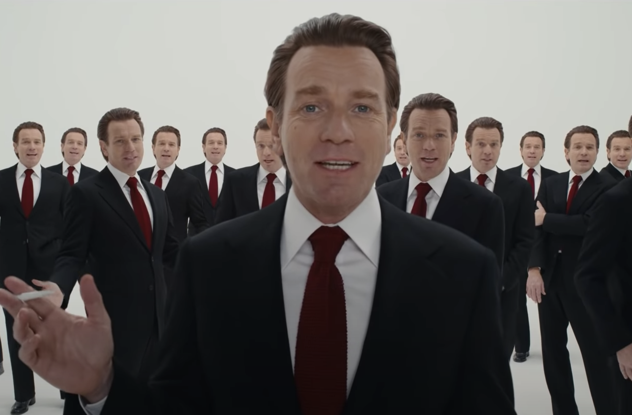 Cena da série Halston. Ewan McGregor, um homem branco, ruivo, de paletó e gravata vermelha está posicionado no centro, da cintura para cima. Atrás, também podemos ver várias reproduções de Ewan, todos usando a mesma roupa.