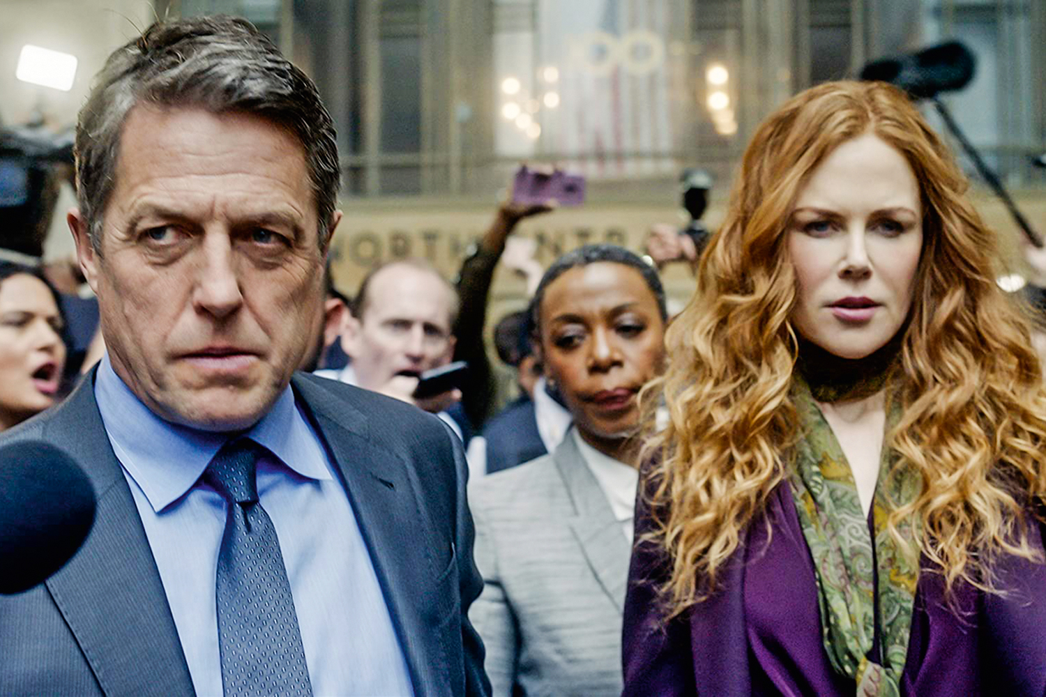 A imagem é uma cena da série The Undoing. Nela Hugh Grant, que interpreta Jonathan Fraser (à esquerda), e Nicole Kidman (à direita), que interpreta Grace Fraser, estão passando entre uma multidão de jornalistas, com um olhar preocupado. Jonathan é um homem branco, com cabelos grisalhos e olhos azuis, ele veste uma camisa, gravata e um terno em tons azuis. Grace é uma mulher branca, de cabelos ruivos e longos, ela veste um vestido e casaco roxos. 