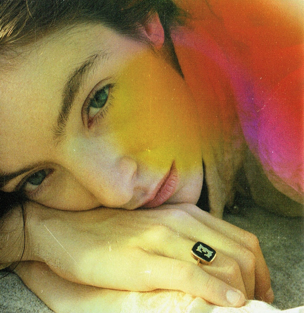 Fotografia quadrada. A câmera está bem próxima do rosto de Lorde, que está apoiado em sua mão. Ela está deitada na areia. Ela usa um anel com a letra E na joia preta. Ela olha diretamente para a câmera. Foi adicionado um efeito de vazamento de luz no canto superior direito, nas cores vermelho, rosa e amarelo, e direção a sua bochecha.