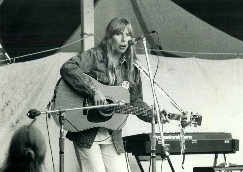 Foto em preto e branco de Joni sentada no chão com um violão no colo. Ela está de cabelo solto e vestido.