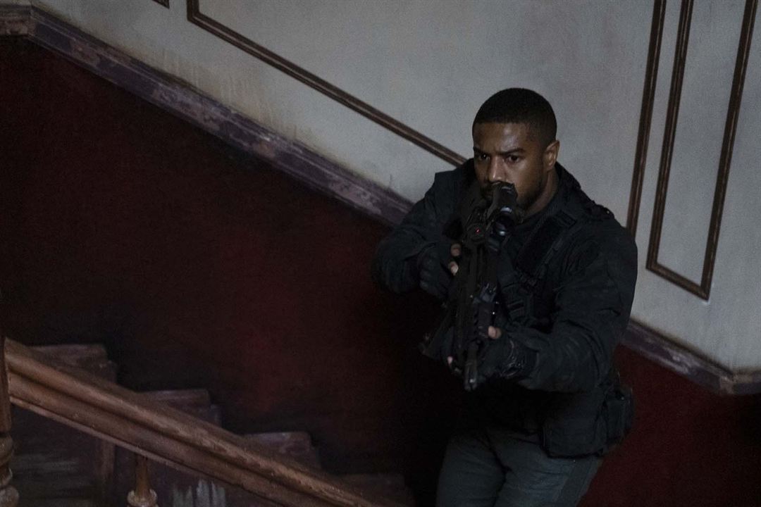Cena do filme Sem Remorso exibe um homem negro, armado e parado num degrau de uma escada desgastada. Ele tem cabeça raspada, tem um olhar desconfiado e veste somente roupas pretas.