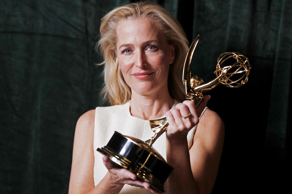Foto de Gillian Anderson. A mulher branca, que tem cabelo loiro e curto, usa um vestido branco. segura na sua frente uma estatueta do Emmy, mostrando para a câmera. A mulher sorri. O fundo é um pano verde.