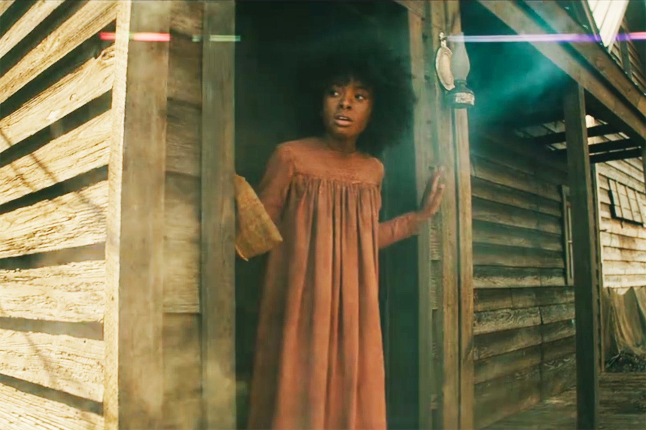 Cena da série The Underground Railroad. A cena mostra Grace, uma criança negra, com vestido vermelho e cabelo black power, olhando para fora, parada em uma porta.