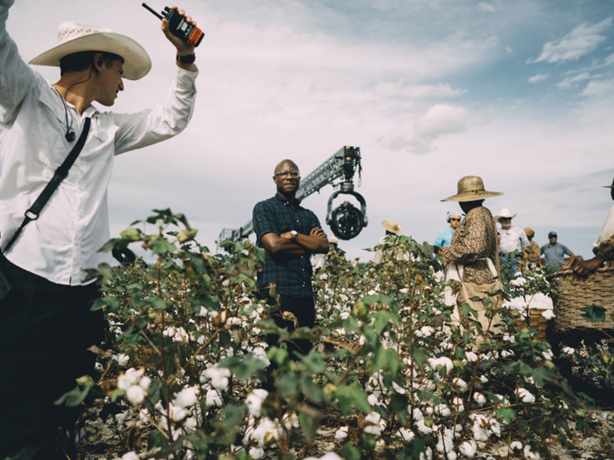 Cena dos bastidores de The Underground Railroad. A foto mostra o diretor Barry Jenkins, um homem negro de óculos, de pé em meio a uma plantação. Ao seu redor, vemos atores e pessoas operando as câmeras.