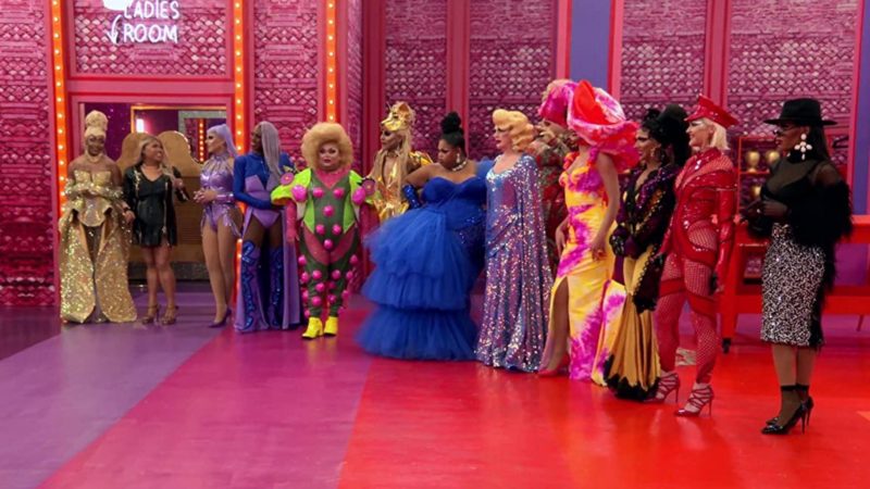 Cena de RuPaul's Drag Race All Stars apresenta as treze participantes montadas e enfileiradas uma ao lado da outra. O ambiente em que elas estão possui paredes rosa, chão listrado de lilás, laranja e magenta e luz de led.