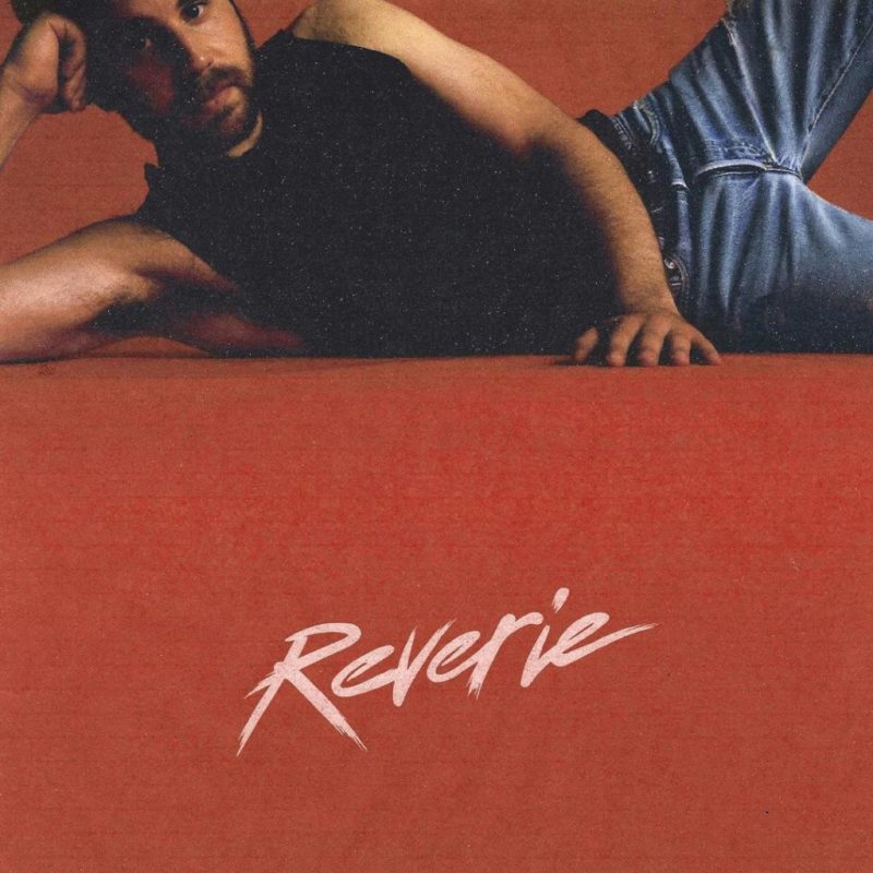 Capa do disco Reverie. Na foto, o artista Ben Platt está deitado na lateral, com a mão apoiando a cabeça. Ele é um homem branco com barba castanha, veste uma camiseta regata preta e uma calça jeans azul. O chão é laranja, e na parte inferior está escrito Reverie em fonte de cor branca.