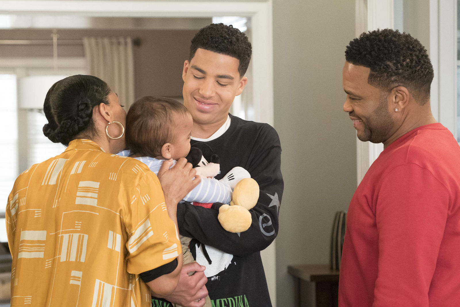 Cena de black-ish. Nela está um garoto negro vestindo blusa preta segurando um bebe no colo, olhando e sorrindo para este. Ao seu lado esquerdo, uma mulher negra com o cabelo preso em tranças e uma blusa amarela beija a cabeça do bebê. Na direita um homem negro de blusa vermelha olha para os três com um sorriso no rosto. 