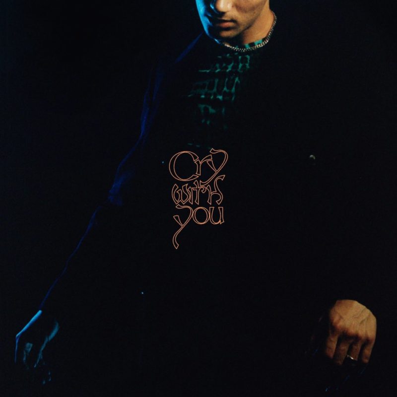 Capa de Cry With You, do Jeremy Zucker. A foto de metade do corpo do cantor mostra a parte de baixo de seu rosto, que é branco, e sua roupa escura, com uma blusa verde por trás da calça e jaqueta preta. No centro da imagem está o nome da música em bege.