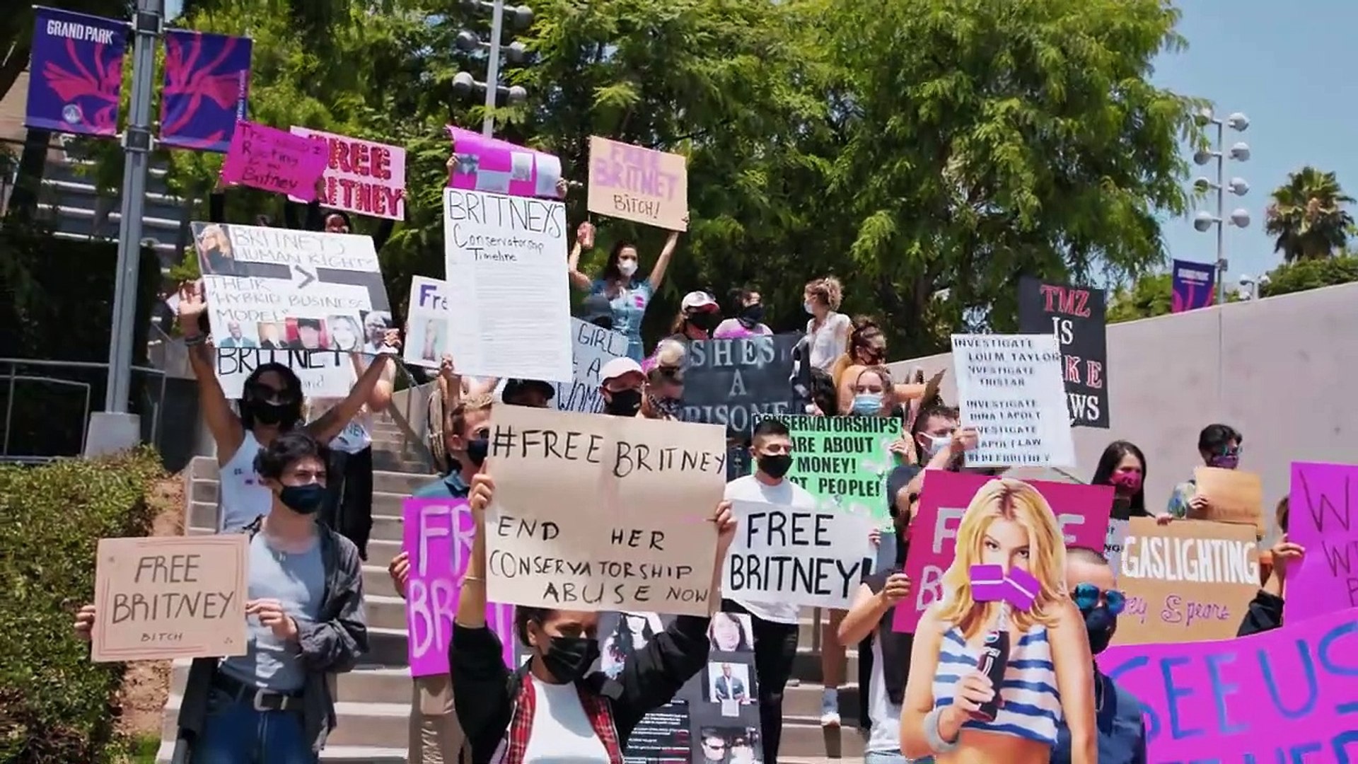 A imagem mostra um protesto a favor da liberdade da cantora Britney Spears, que está judicialmente dependente de seu pai em uma conservadoria. A foto é uma cena do documentário Framing Britney Spears, e mostra diversas pessoas na calçada de uma rua com cartazes pedindo a liberdade da cantora.
