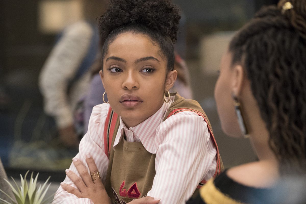 Cena de grown-ish. Nela está a foto de uma mulher negra, de cabelo cacheado preso em um coque alto. Ela veste uma regata marrom por cima de uma camisa branca e rosa listrada, com um conjunto de brincos de argolas douradas. Ela olha para uma garota na sua frente, que só é possível ver parte das costas e do cabelo. O fundo está desfocado.