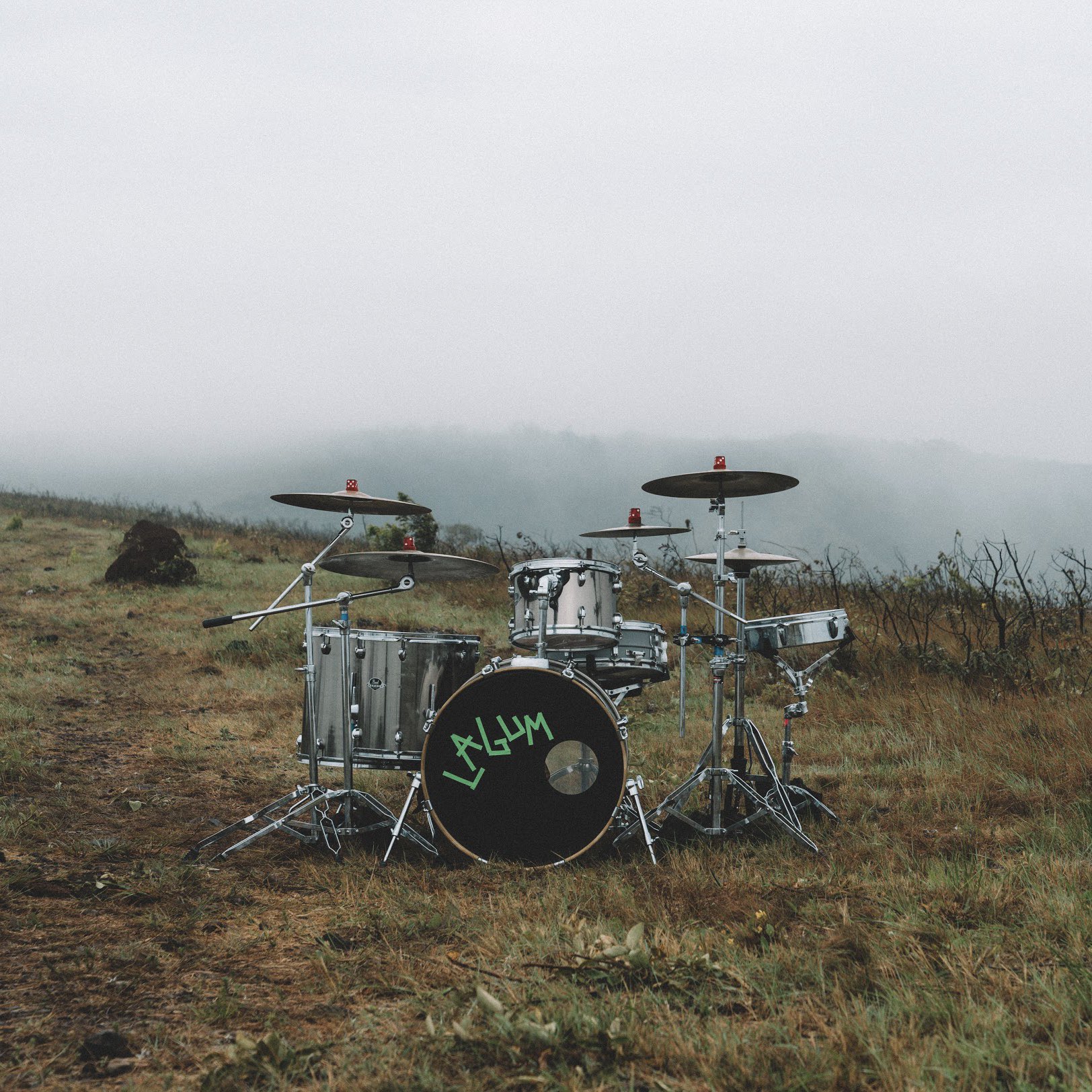 Texto alternativo: Fotografia da bateria da Lagum num cenário de campo. Existe uma vegetação rasteira com neblina ao fundo. Na frente, existe uma bateria prata com o tambor preto escrito Lagum com letra verde.