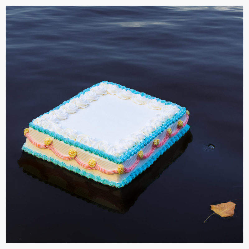 Capa do álbum Sunny Bay de Odd Beholder. Imagem quadrada com bordas laterais brancas. Fotografia de um bolo de aniversário flutuando sobre a água. O bolo é confeitado com glacê branco, azul, rosa e amarelo. Também sobre a água está uma folha seca ocre, no canto inferior direito.