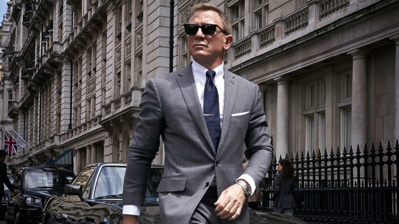 Cena do filme 007 - Sem Tempo Para Morrer exibe um homem branco, de meia idade. Ele veste um terno cinza, com gravata azul-escura. Tem cabelo curto, loiro, e usa óculos escuros. Ao fundo, vemos carros estacionados em uma rua de Londres. 
