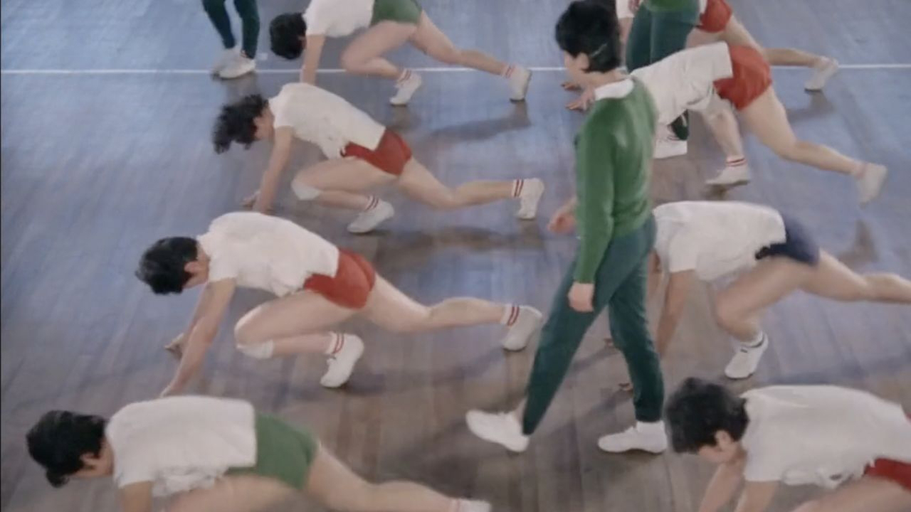 Cena do documentário As Bruxas do Oriente. A imagem mostra um time de vôlei feminino do Japão treinando. Duas fileiras de mulheres uniformizadas com camisetas brancas e shorts vermelhos estão fazendo flexões no chão. No meio das fileiras, existe um homem, o treinador, que usa um moletom verde e observa as atletas.
