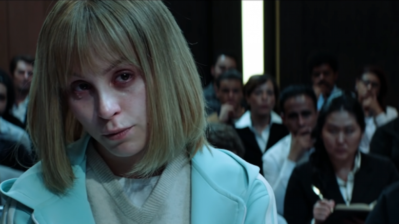 Cena do filme A Menina que Matou os Pais. Nela vemos Carla Diaz no banco do tribunal. Ela é branca, loira e usa uma blusa azul bebê.