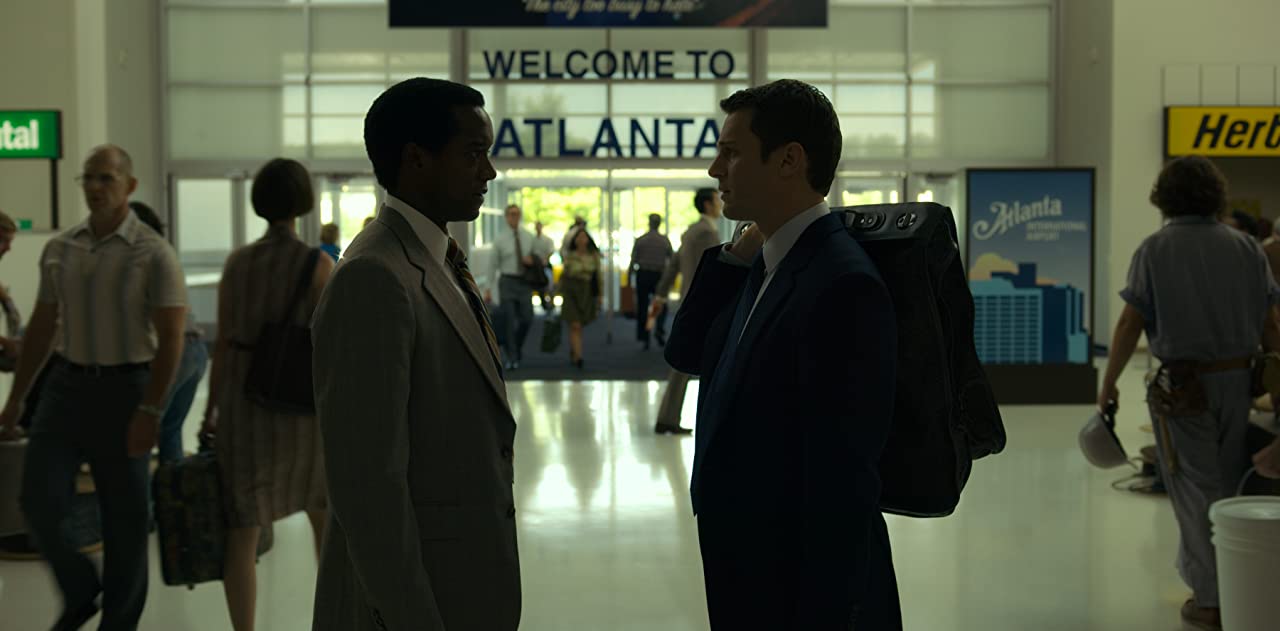 Cena da série Mindhunter. Na imagem, Holden e Jim se encaram frente a frente na saída de um aeroporto. Jim está à esquerda e é um homem negro de paletó cinza. Holden está à direita e é um homem branco de paletó preto segurando uma mala com a mão direita no ombro. No centro e ao fundo está escrito “Bem vindo à Atlanta” em inglês.