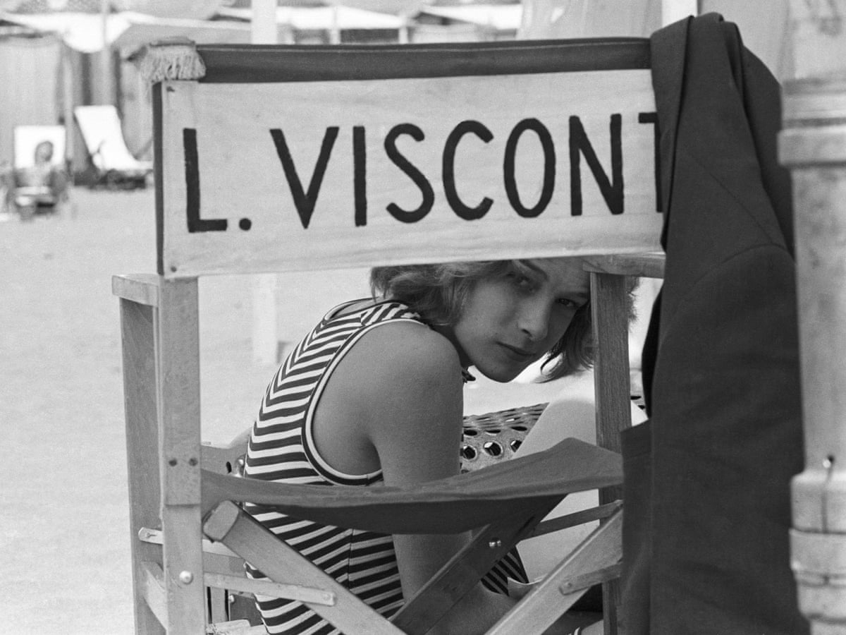 Cena do documentário O Garoto Mais Bonito do Mundo. Em preto e branco, mostra um garoto branco e loiro escondido atrás de uma cadeira de set de gravações. Nas costas da cadeira, está escrito L. Visconti, o nome do diretor. 