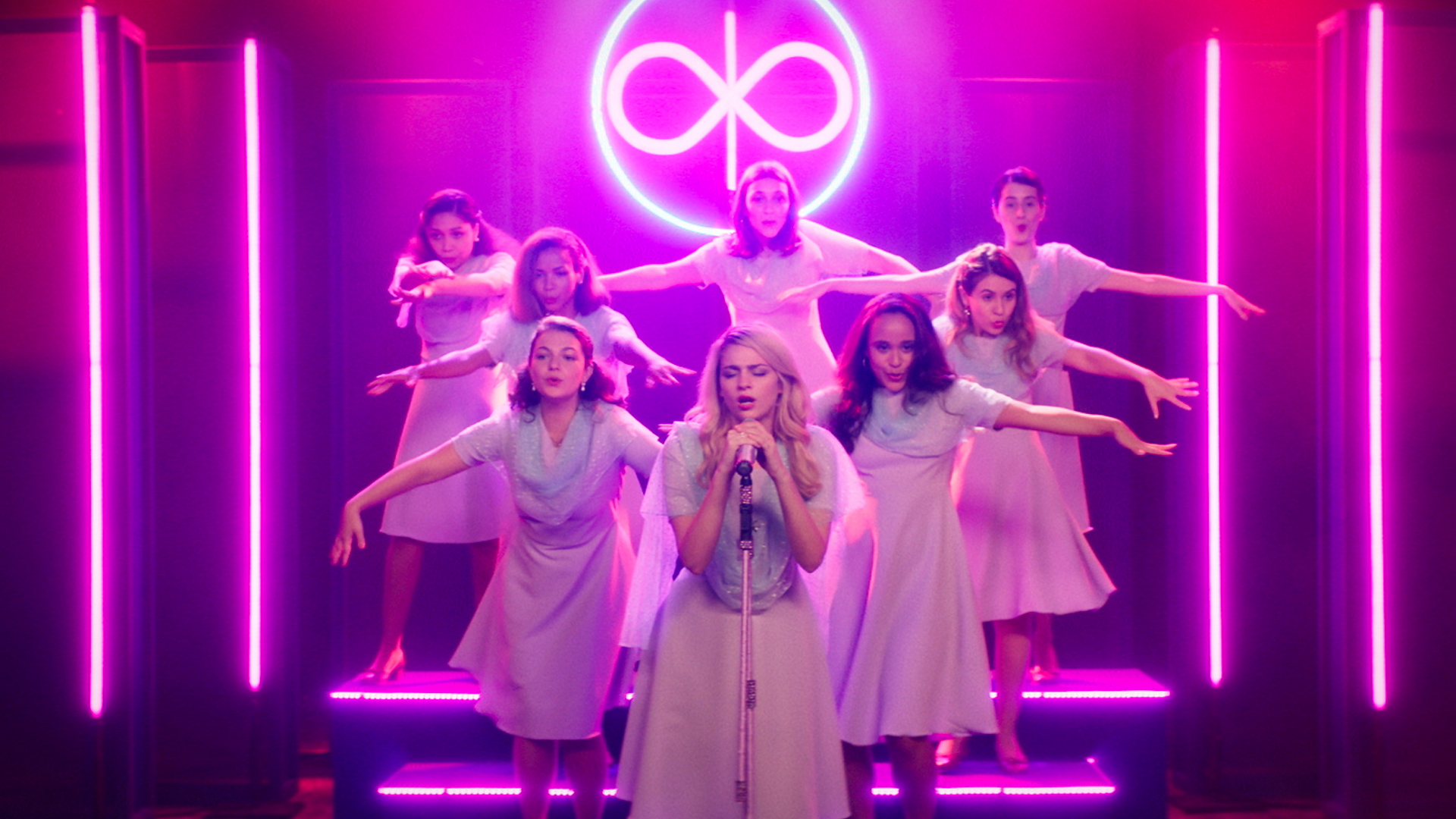 Cena do filme Medusa. 8 mulheres estão em cima de um palco rosa neon com vestidos brancos. A da frente, loira, está cantando no microfone. As outras coreografam ao fundo.