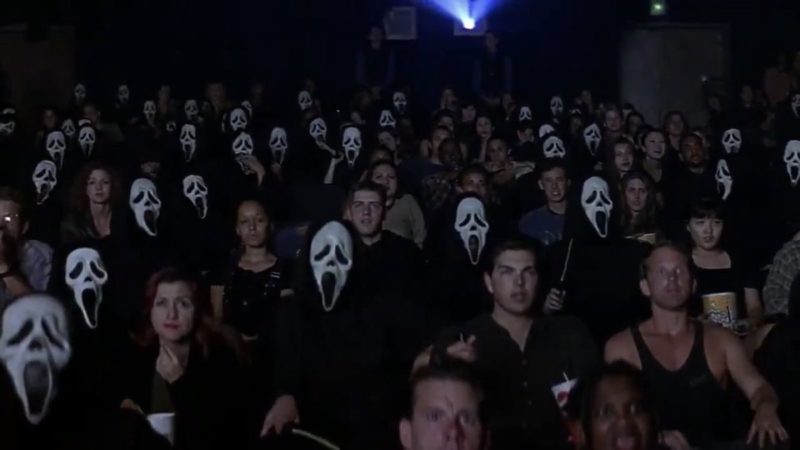 Cena do filme Pânico. Nela vemos uma sala de cinema lotada. A maioria das pessoas usa a máscara branca com olhos pretos e a boca aberta.