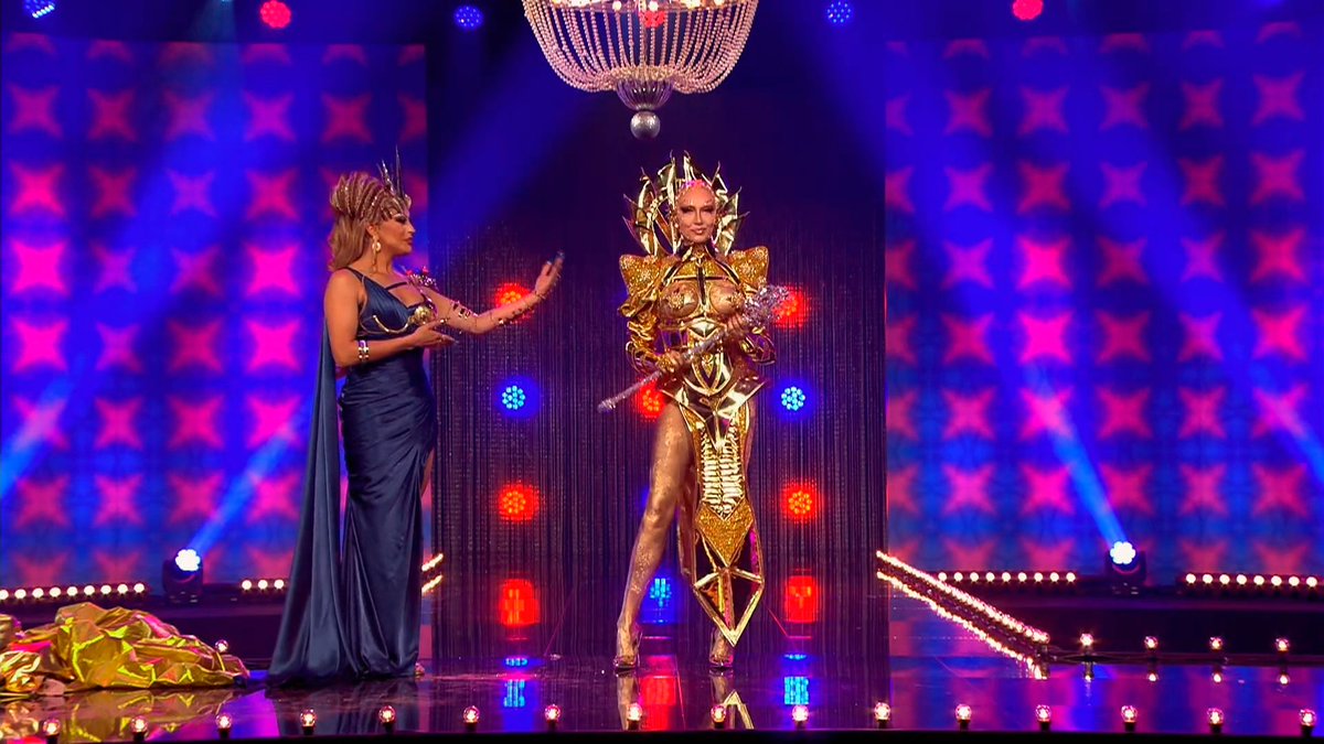 Cena de Drag Race Holland. A cena mostra Envy Peru coroando Vanessa van Cartier, que usa uma roupa dourada e segura um cetro prateado nas mãos. 