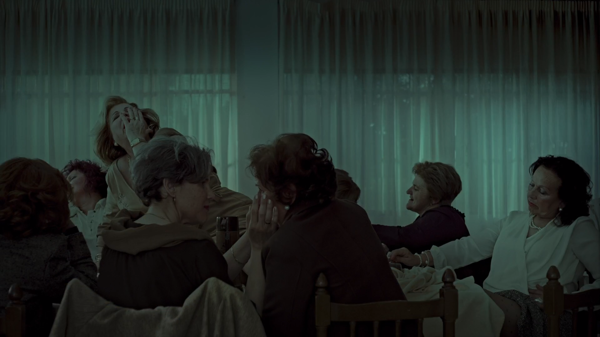 Cena do filme Um Forte Clarão exibe várias mulheres brancas idosas ao redor de uma mesa. Elas parecem estar sob efeito de alguma droga. Algumas mulheres tocam no rosto das outras e, em cima da mesa, uma mulher está deitada com a mão no rosto. Parece extasiada. 