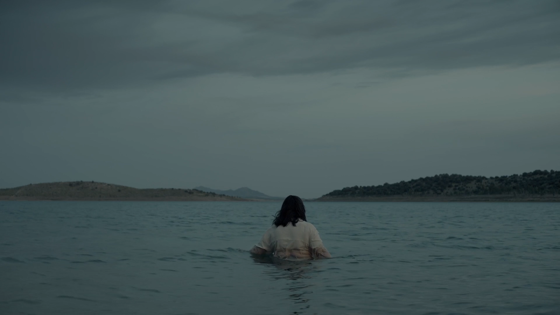 Cena do filme Um Forte Clarão exibe uma mulher parada no mar. A água alcança seu umbigo e ela está de costas para a câmera. Veste uma camiseta branca e tem cabelo preto, na altura dos ombros. Ao longe, vemos duas ilhas. 