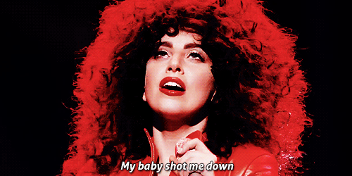Cena do show Cheek To Cheek Live from Jazz At Lincoln Center. GIF retangular. A cantora Lady Gaga ocupa quase todo o GIF. Ela é uma mulher branca, de batom vermelho e usa uma peruca que imita os cabelos da também cantora Cher. Olhando para cima, Gaga abaixa o microfone que segura em uma das mãos. No meio da parte inferior, lemos “my baby shot me down”, um trecho da letra da canção Bang Bang (My Baby Shot Me Down).