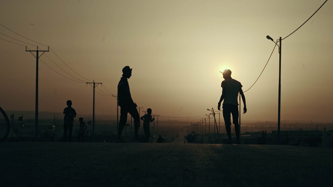 Imagem retangular retirada do filme Capitães de Zaatari. O cenário é o pôr-do-sol de uma estrada de terra. Vemos nas extremidades vários postes elétricos e, no centro, a silhueta de alguns garotos jogando futebol na rua.
