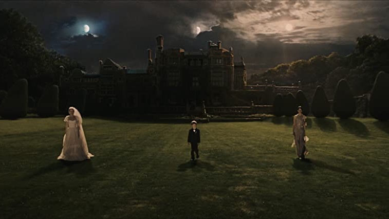 Cena do filme Melancolia em que há uma mulher vestida de noiva à esquerda, uma criança de terno preto no centro e uma mulher de vestido cinza à direita. Os três estão em um gramado e ao fundo há uma mansão.