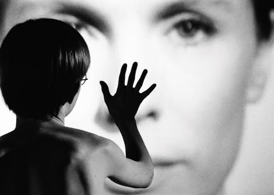 Cena do filme Persona apresenta uma imagem em branco e preto de um menino de costas com a mão direita erguida sobre a foto de uma mulher branca, que toma conta de todo o plano.