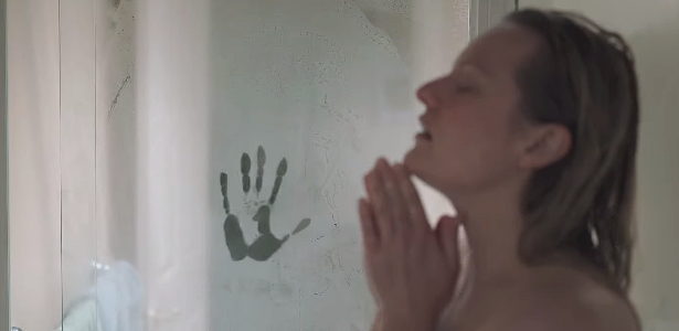 Cena do filme O Homem Invisível. A personagem Cecília (Elisabeth Moss), de cabelos loiros, está de lado num box de banheiro, enquanto no segundo plano o foco está na mão do Homem Invisível marcada no vidro do box embaçado.