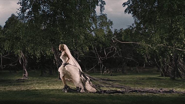 Cena do filme Melancolia que apresenta uma mulher branca, loira, vestida de noiva correndo. Há diversas raízes que a prendem pela perna, dificultando sua fuga. No fundo da imagem aparecem mais árvores com cipó.
