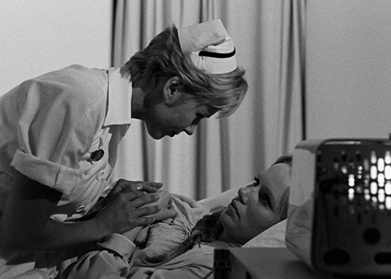 Cena do filme Persona apresenta uma imagem em branco e preto de uma enfermeira branca de cabelos curtos loiros debruçada sobre uma paciente. Esta é uma mulher branca de cabelos longos, que está deitada em uma cama de hospital.