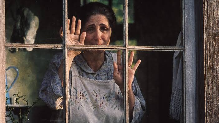 Cena do filme Lavoura Arcaica no qual há uma mulher branca de meia idade, de cabelos castanhos presos, com um vestido e um avental, olhando através da janela enquanto chora.