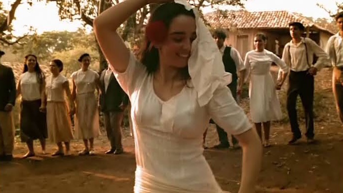 Cena do filme Lavoura Arcaica mostra uma mulher branca jovem, de cabelos castanhos, que usa um vestido branco enquanto dança. Ao fundo da imagem há outras pessoas lado a lado.