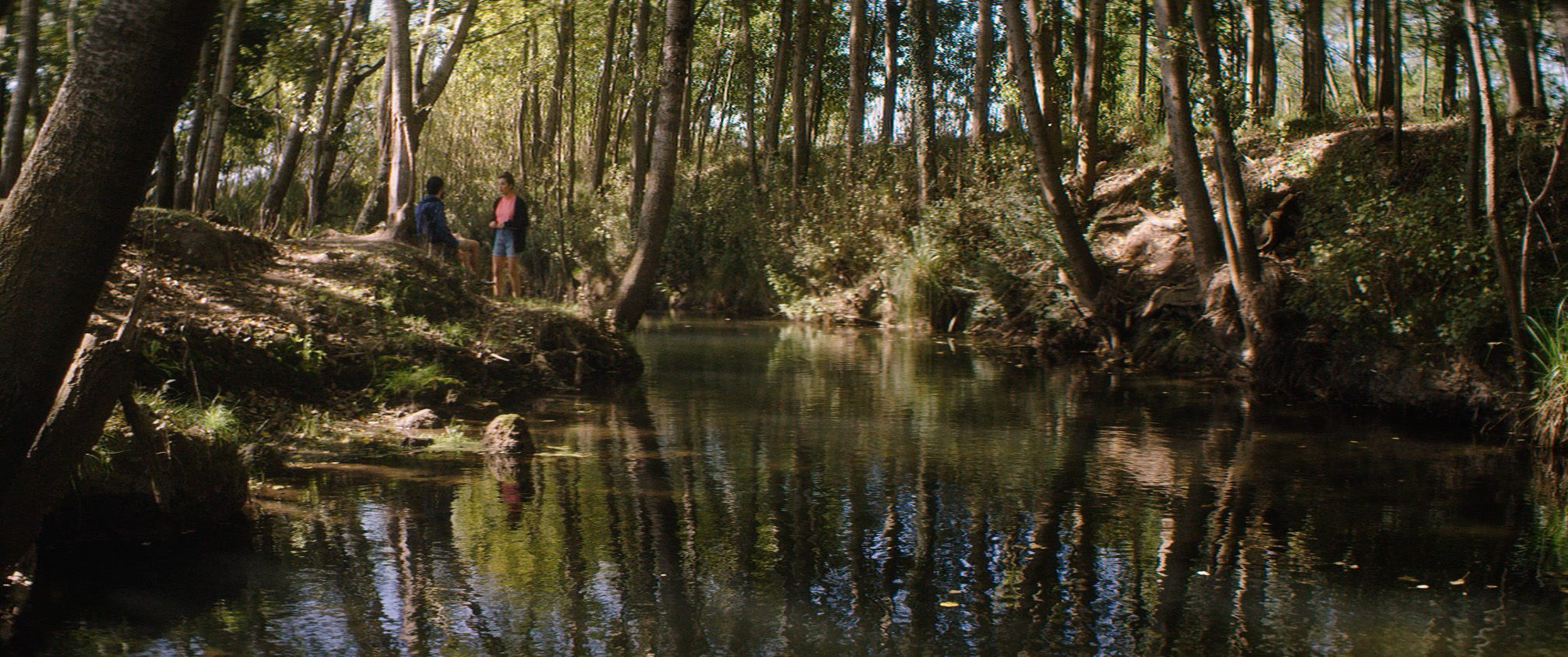Cena do filme Os Inventados. A foto é uma paisagem de uma floresta, ensolarada, com um lago no centro. No canto, podemos ver de longe dois personagens de pé, um de frente para o outro.