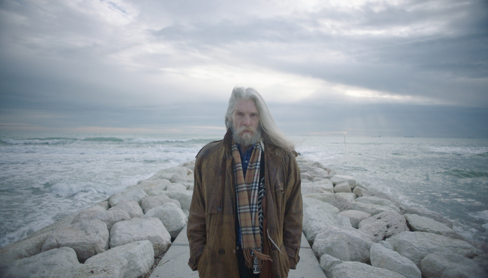 Cena do documentário O Garoto Mais Bonito do Mundo. Mostra um homem idoso, de cabelos brancos e barba branca, compridos, parado próximo ao mar. Ao fundo, vemos a água azul.