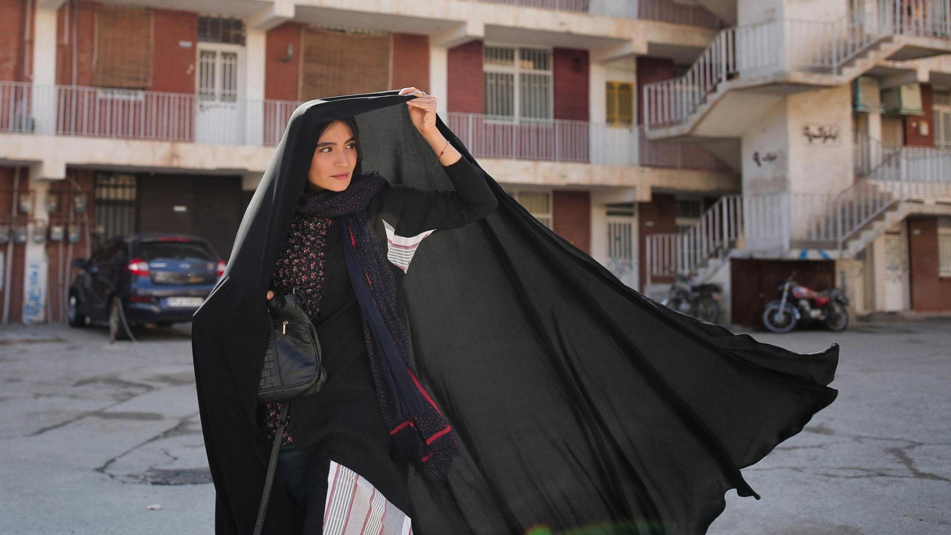 Cena do filme Um Herói. Mostra uma mulher iraniana usando vestimentas pretas e cobrindo sua cabeça com um tecido da mesma cor. Ao fundo, vemos casas de tijolo vermelho. 