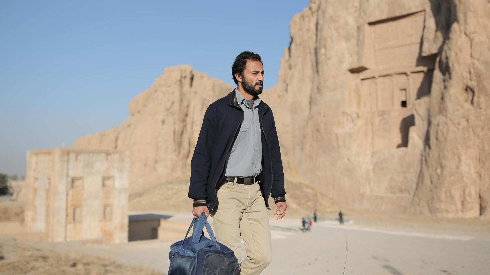 Cena do filme Um Herói. Mostra um homem adulto, iraniano, caminhando perto de uma montanha clara. Está de dia e está bem sol, e ele segura uma mala azul com a mão direita. 