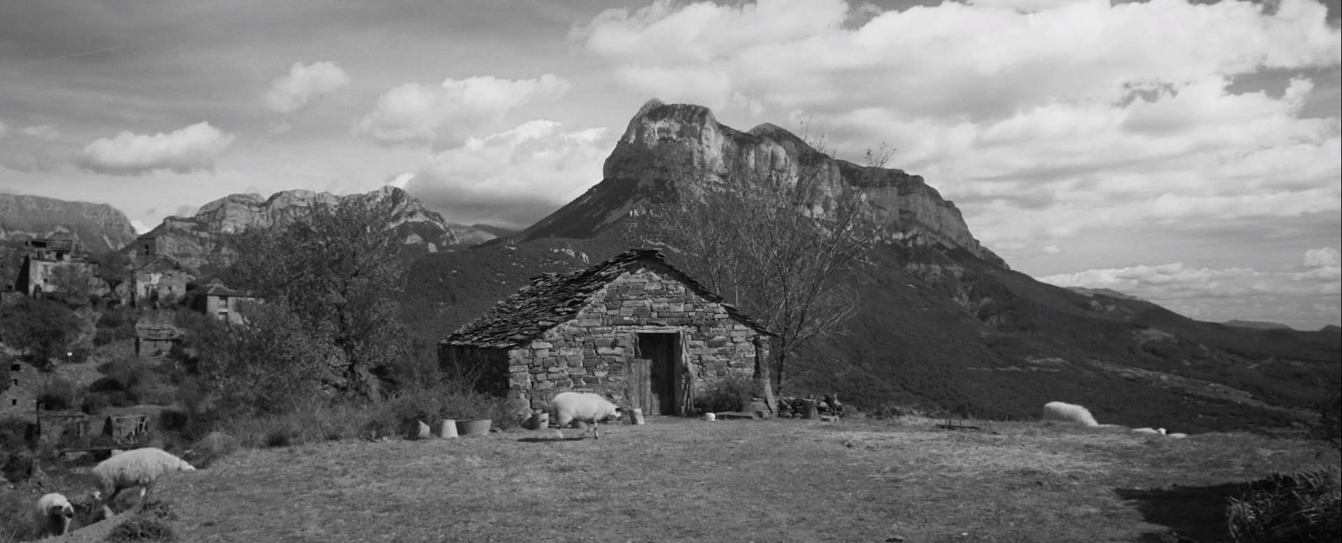 Cena do filme Armugan, em preto e branco. Vemos uma cabana em uma região isolada, nas montanhas. Em frente dela, quatro carneiros se alimentam da vegetação no solo. Ao fundo, vemos algumas casas.