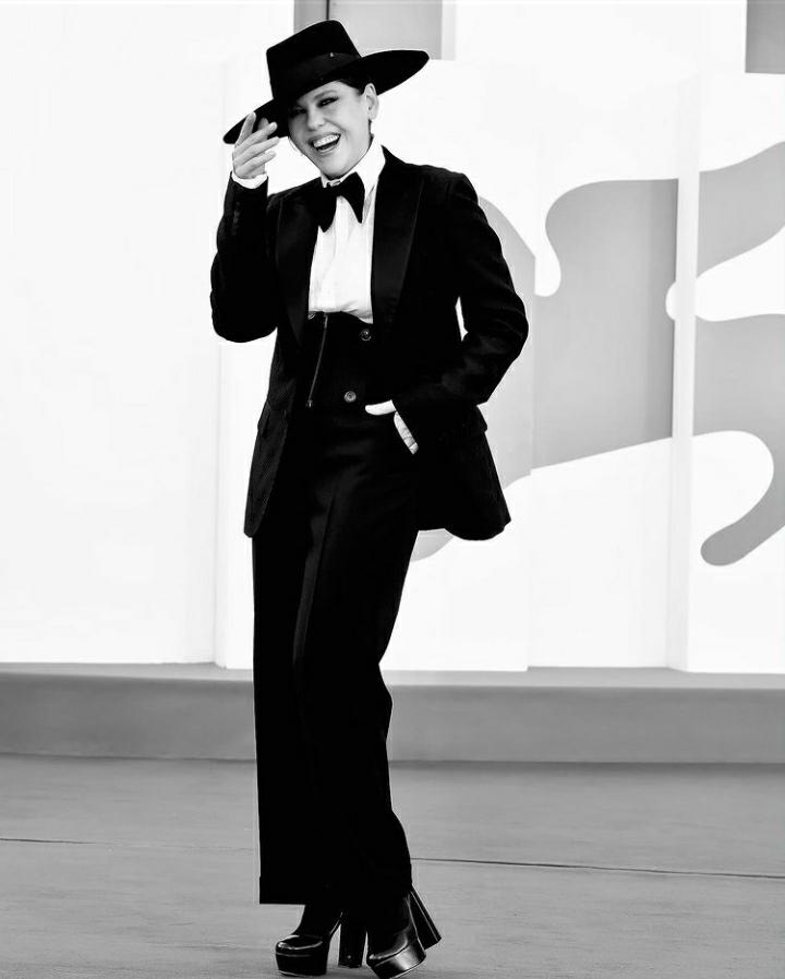 Foto em preto e branco. Bárbara Paz de chapéu e paletó pretos, gravata borboleta, calça e botas pretas. Com o fundo do festival.