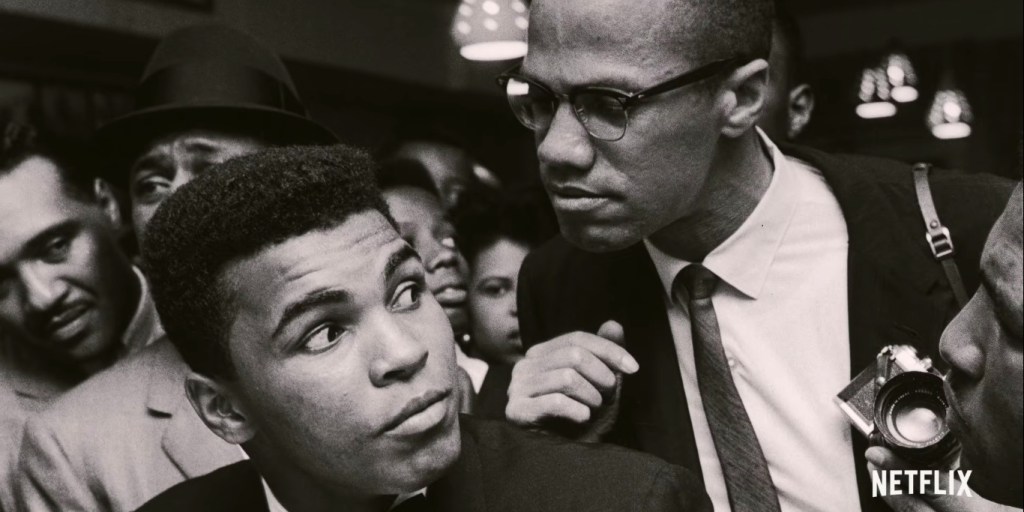 Cena do documentário Irmãos de Sangue. A imagem é preto e branca e Muhammad Ali está sentado com o pescoço virado para o lado, ouvindo Malcolm X que está atrás dele. Ali é um homem negro de cabelo crespo, está de terno preto e sua expressão é de curiosidade. Malcolm é um homem negro um pouco mais velho, com os cabelos cortados curtos e óculos, ele usa terno, camisa e gravata, e uma de suas mãos apoia no ombro de Ali, enquanto a outra segura uma pequena câmera; sua expressão é séria e ele está olhando para Ali. No fundo há diversos homens.
