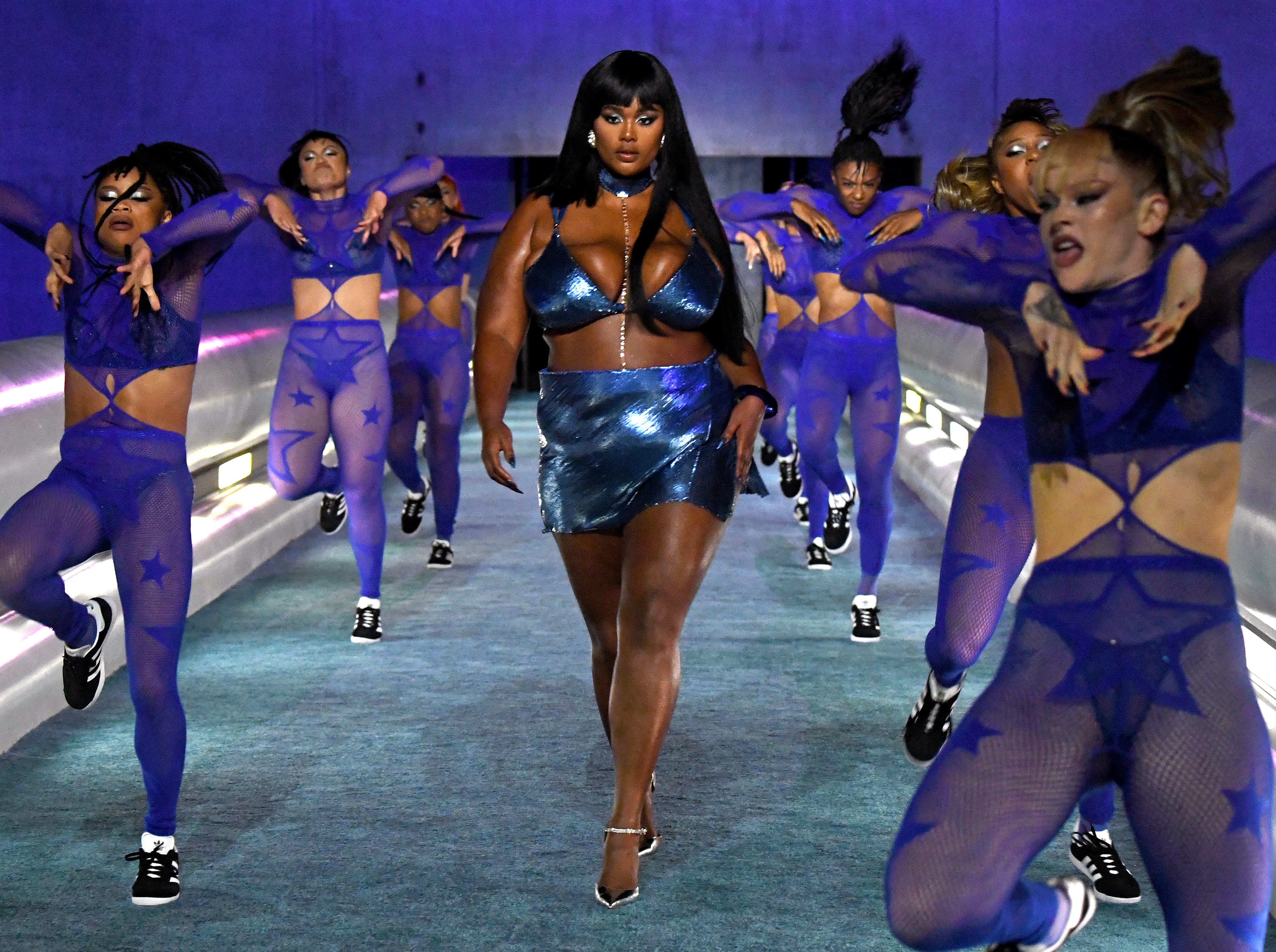 Cena do Fenty X Savage Show. Uma mulher negra, gorda, de cabelos pretos e grandes, usando um sutiã e saia azuis metálicos desfilando entre dez dançarinas, usando macacões azuis. Ao lado delas há corrimões e luzes azuis ao fundo.