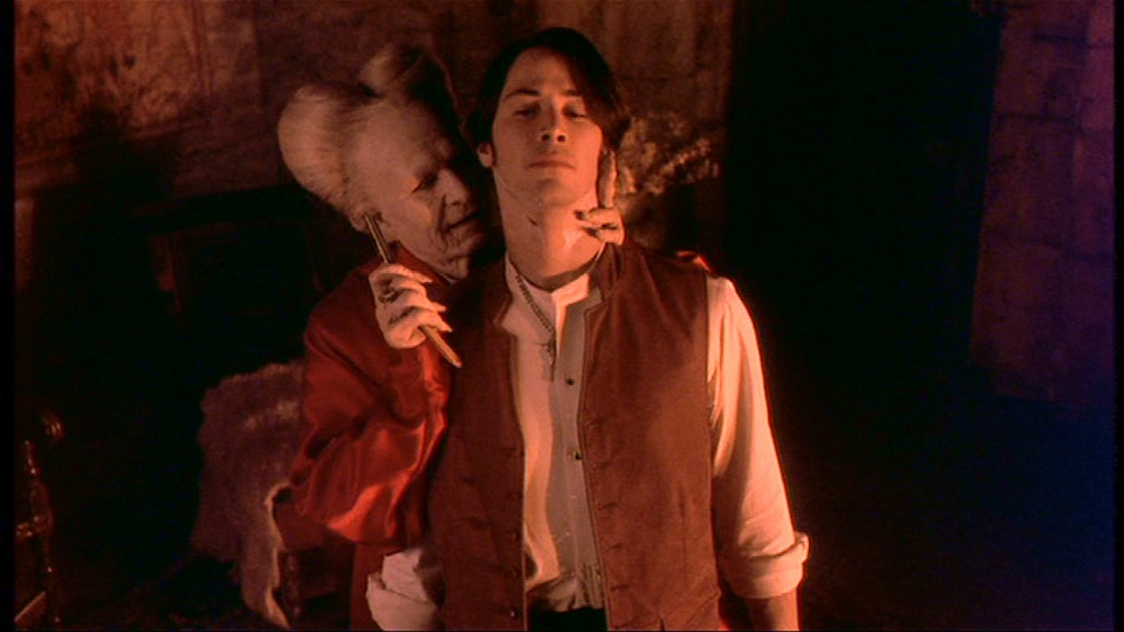 Cena do filme Drácula de Bram Stoker de 1992. Na imagem estão o Conde Drácula interpretado por Gary Oldman, e a sua esquerda Jonathan Harker interpretado por Keanu Reeves. O vampiro tem a pele branca pálida, unhas compridas e cabelos brancos, veste uma camisa de cetim vermelha e segura um objeto pontiagudo próximo ao pescoço de Jonathan. Harker por sua vez veste uma camisa branca e um colete marrom desabotoado, tem pele branca e cabelos escuros.